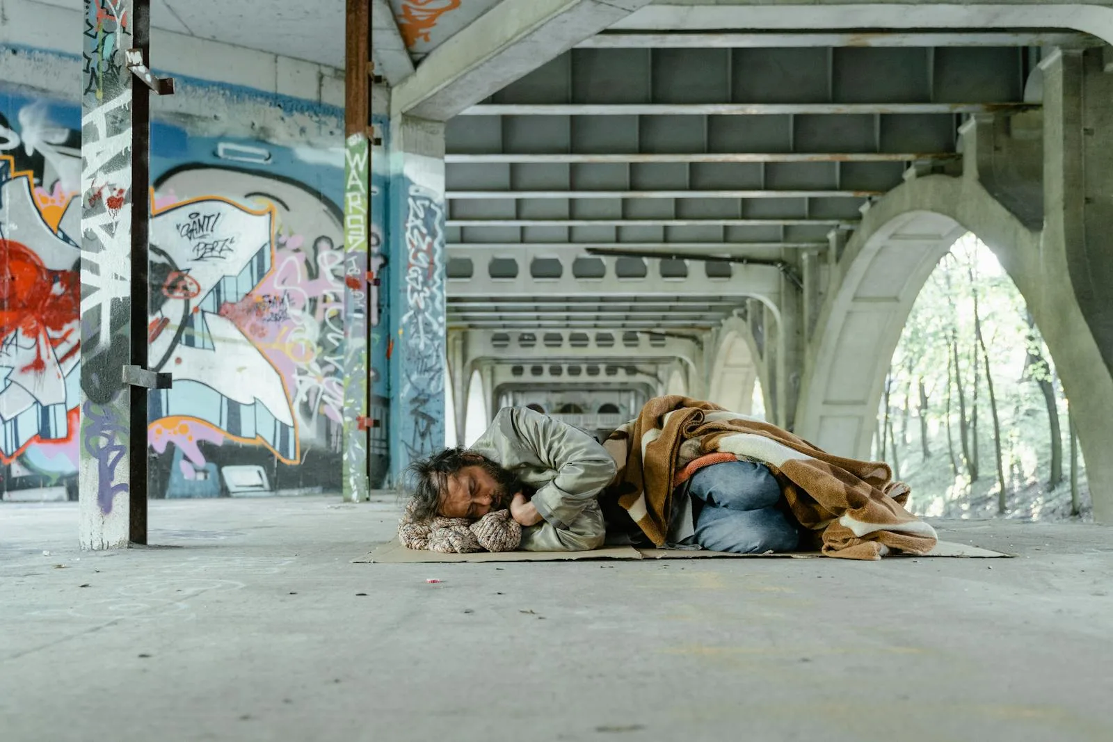 A homeless man sleeping on cardboard under a graffiti-laden bridge.