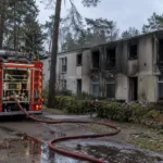 Wóz strażacki przed osmalonym blokiem mieszkalnym ośrodka wśród sosen, wąż strażacki na mokrej ziemi