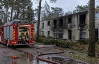 Wóz strażacki przed osmalonym blokiem mieszkalnym ośrodka wśród sosen, wąż strażacki na mokrej ziemi