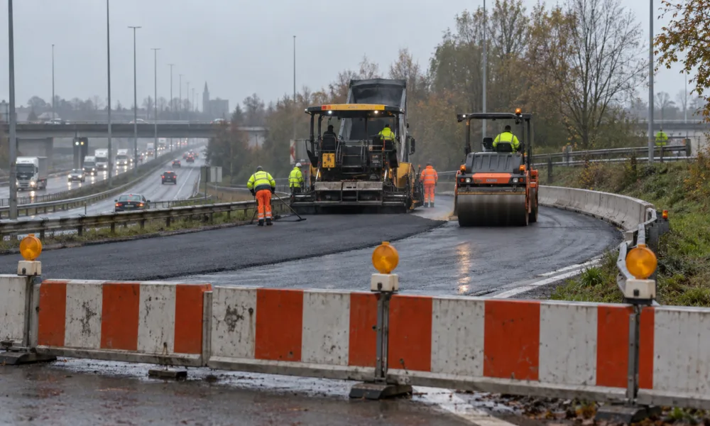 Zamknięty wjazd na obwodnicę pod Gandawą, biało-czerwone zapory i maszyny kładące nowy asfalt w pochmurny październikowy dzień