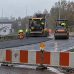 Zamknięty wjazd na obwodnicę pod Gandawą, biało-czerwone zapory i maszyny kładące nowy asfalt w pochmurny październikowy dzień