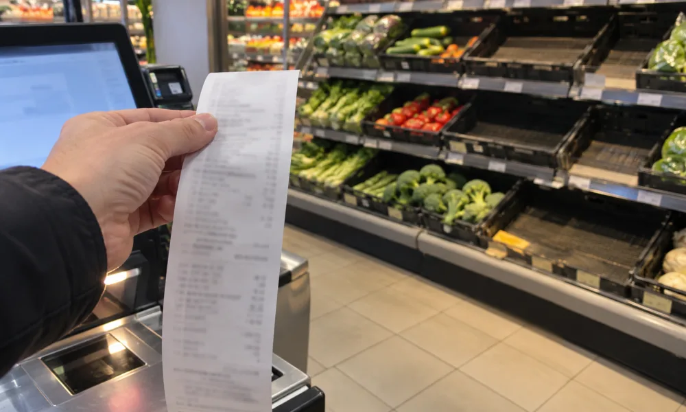 Dłoń trzymająca długi paragon przy kasie samoobsługowej, w tle półki z warzywami i puste skrzynki