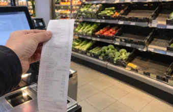 Dłoń trzymająca długi paragon przy kasie samoobsługowej, w tle półki z warzywami i puste skrzynki