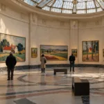 Sala wystawowa w stylu art déco z obrazami belgijskich pejzaży i zwiedzającymi widzianymi z tyłu
