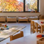 Jasna sala żłobka we Flandrii z niskimi stolikami, klockami na dywanie i rzędem małych łóżeczek pod oknem