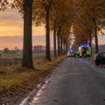 Wiejska droga obsadzona drzewami we Flandrii Zachodniej, rozbity samochód przy drzewie i służby ratunkowe - ilustracja SI