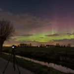 Zielono-różowa zorza polarna nad płaskimi polami we Flandrii, statyw z aparatem przy wiejskiej drodze