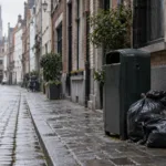 Worki ze śmieciami i karton zostawione przy koszu na wąskiej brukowanej uliczce w Antwerpii, w tle sprzątacz miejski