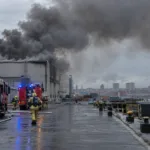 Gęsty dym nad halą przemysłową w porcie, wozy strażackie na nabrzeżu