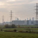 Słupy wysokiego napięcia na łąkach przed halami fabrycznymi i instalacjami chemicznymi we Flandrii, w tle turbiny wiatrowe