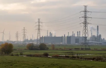 Słupy wysokiego napięcia na łąkach przed halami fabrycznymi i instalacjami chemicznymi we Flandrii, w tle turbiny wiatrowe