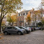 Niewielki plac otoczony ceglanymi kamienicami, środek zajęty przez zaparkowane samochody, kilka drzew