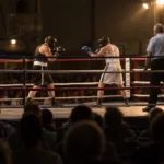 Dwóch bokserów w kaskach i sędzia na ringu w hali sportowej, na pierwszym planie widownia