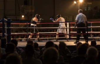 Dwóch bokserów w kaskach i sędzia na ringu w hali sportowej, na pierwszym planie widownia