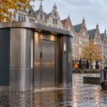 Nowoczesna toaleta publiczna na placu w Antwerpii, w tle kamienice i wieża katedry