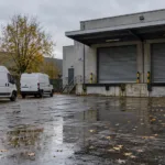 Pusty plac firmy logistycznej z zamkniętymi bramami rampy załadunkowej i dwiema białymi furgonetkami w deszczowy jesienny dzień