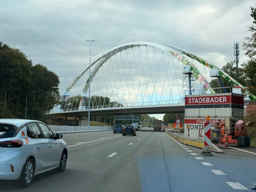 Nowy biały most łukowy nad autostradą E40 w Bertem, pod nim jadą samochody, z prawej znak ograniczenia prędkości do 50 km/h i kontenery wykonawcy