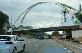 Nowy stalowy most łukowy z malowidłem kwiatów nad autostradą E40 w Bertem, widziany z jezdni, pod mostem ruch samochodów