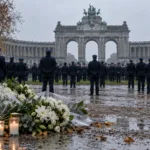 Policjanci w mundurach przed łukiem w parku Cinquantenaire, kwiaty i znicze na pierwszym planie
